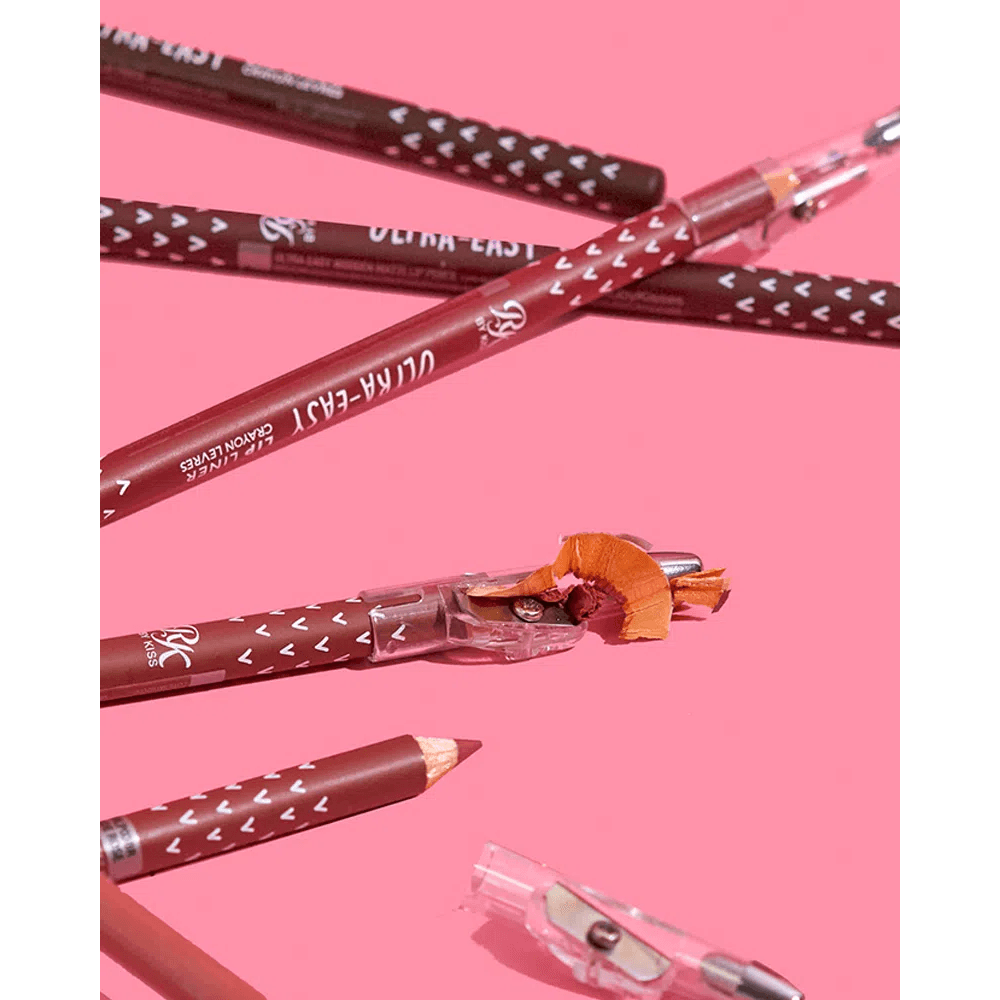 Lápis RK by Kiss Pale Nude sendo apontado, com aparas de madeira visíveis em fundo rosa.