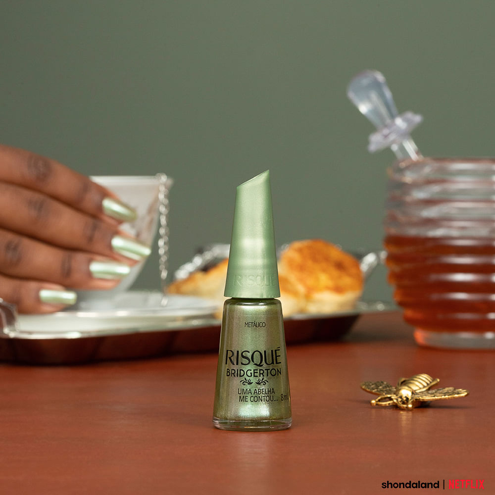 Frasco de esmalte Risqué Bridgerton verde metálico em cena com mão segurando xícara