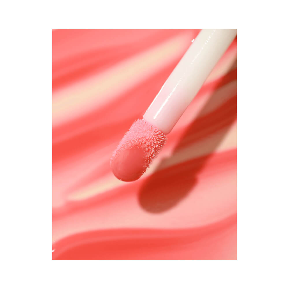 Aplicador de gloss labial, aplicado sobre uma superfície rosa claro, deixando um traço rosa claro. O fundo é branco.