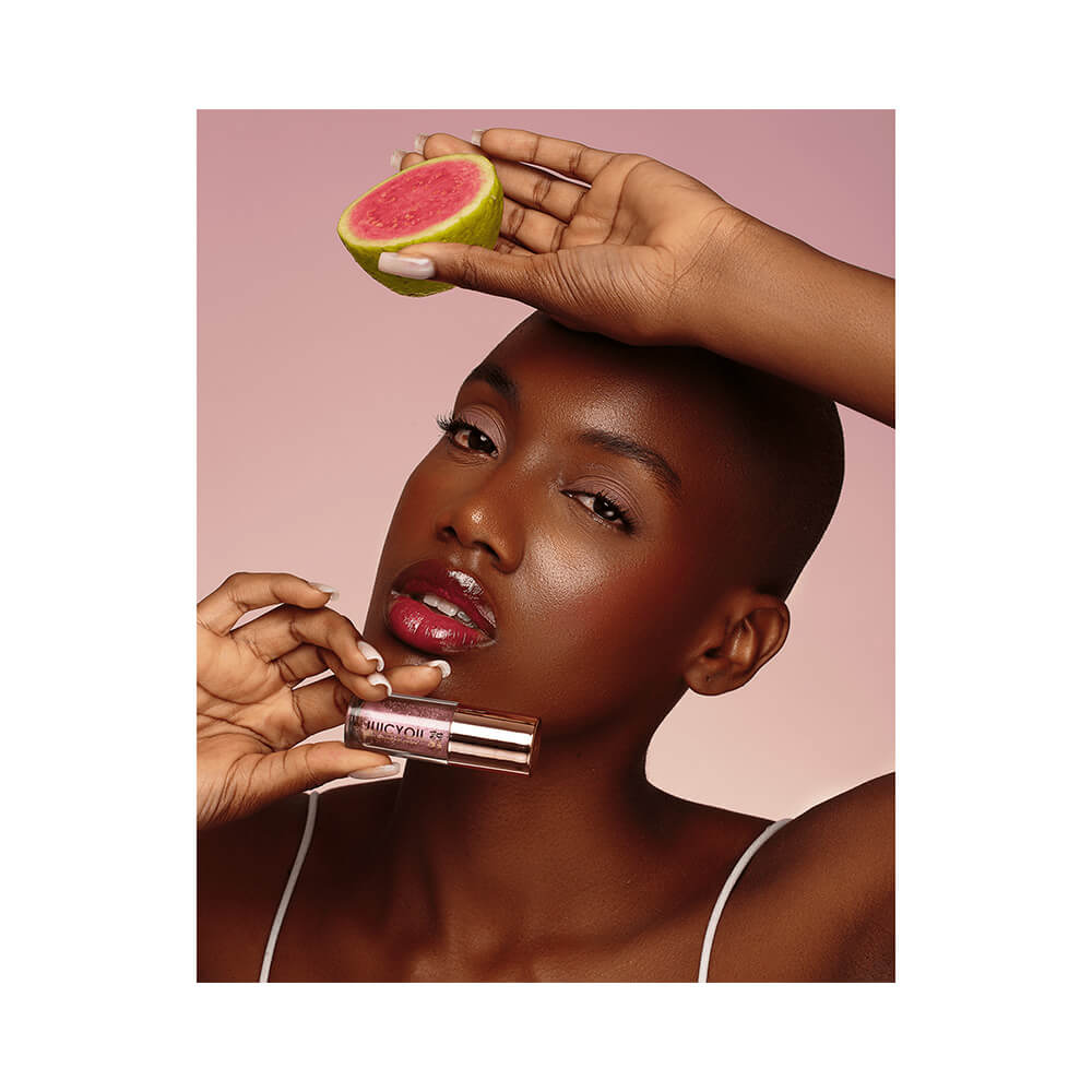 Mulher aplicando batom Juicy Oil Bruna Tavares Guava segurando uma fatia de goiaba