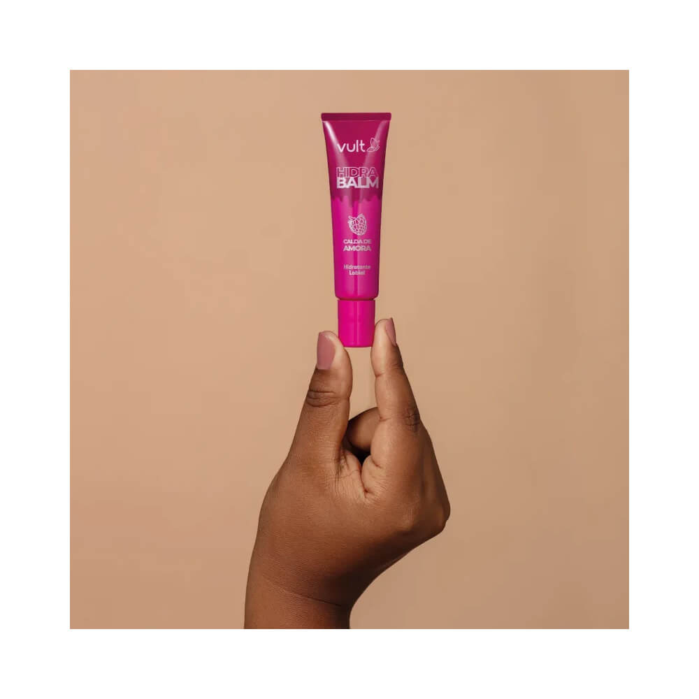 Mão segurando o tubo de hidratante labial Vult Hidra Balm Calda de Amora sobre fundo bege