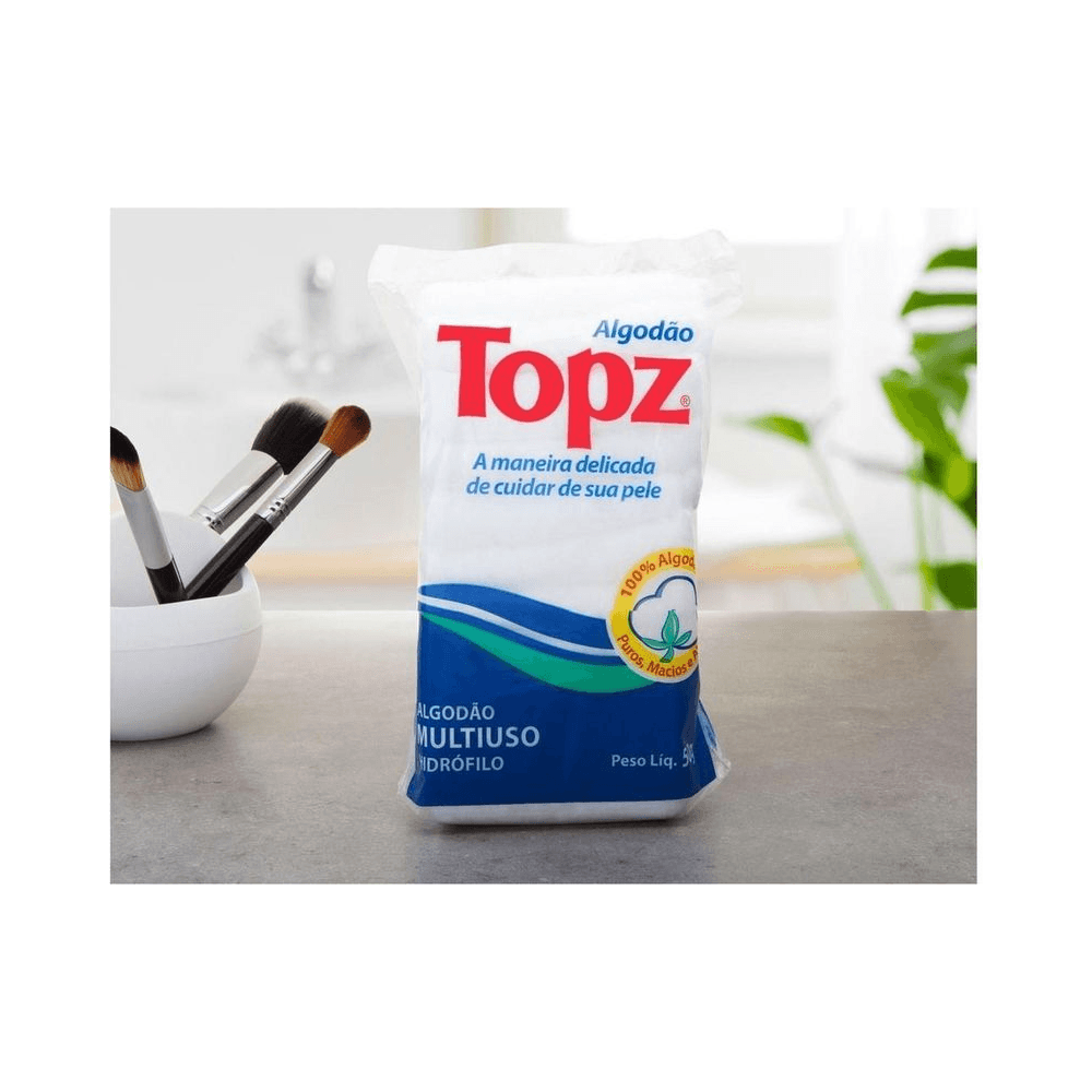 Pacote de Algodão Topz Multiuso 50g sobre superfície com pincéis ao fundo, destacando selo 100% algodão