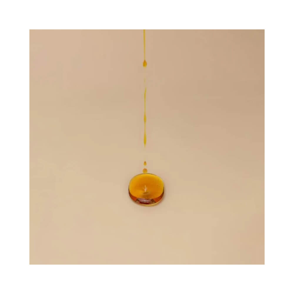 Gotas de óleo âmbar caindo sobre superfície bege, ilustrando textura hidratante do spray bronzeador