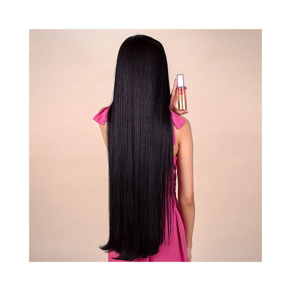 Uma mulher com cabelos longos e pretos segurando um frasco de óleo capilar Forever Liss Cresce e Aparece em cor rosa, com tampa branca, contra um fundo rosa claro.