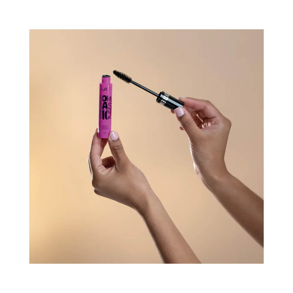 Mão segurando a Máscara de Cílios Vult Extreme Bombastic! com aplicador preto e frasco rosa em fundo amarelo.