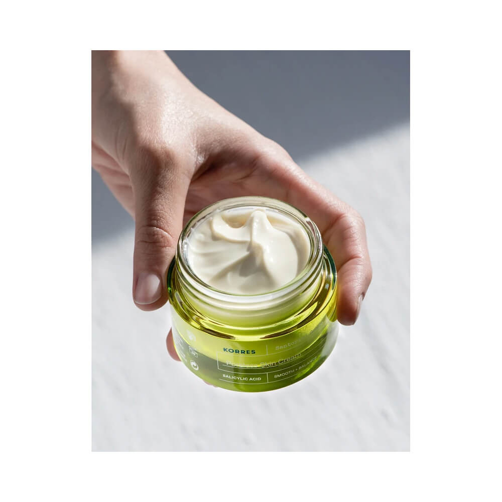 Mão segurando o frasco aberto do Gel Creme Korres Santorini Grape mostrando o creme branco e cremoso