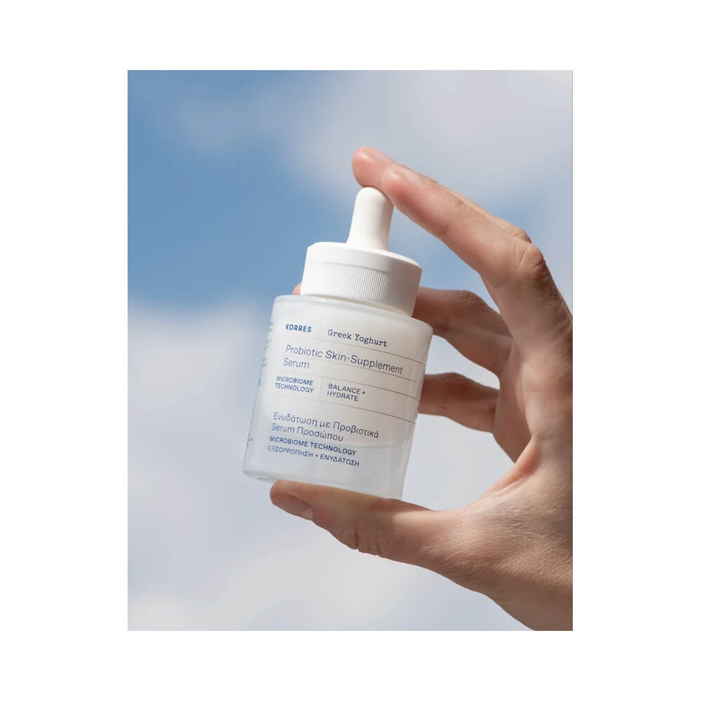 Mão segurando frasco do Sérum Facial Korres Greek Yoghurt Hidratação Ativa 30ml com fundo azul e nuvens