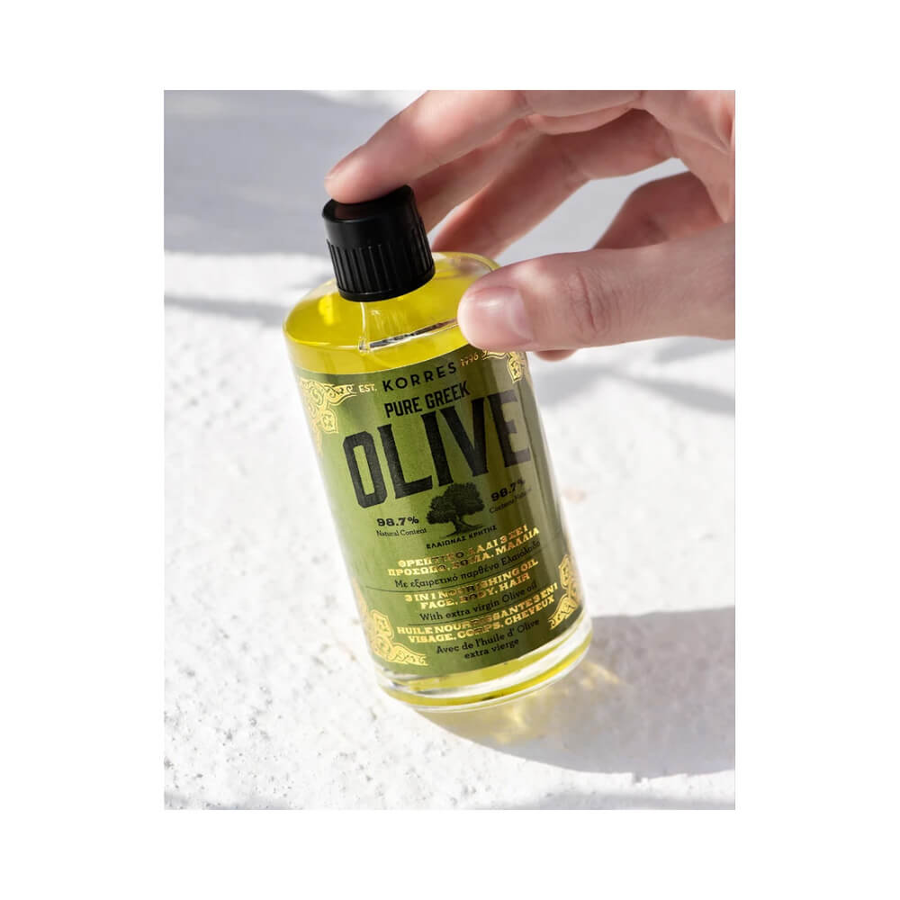 Mão segurando frasco transparente de óleo facial amarelo claro Korres Pure Greek Olive 3 em 1