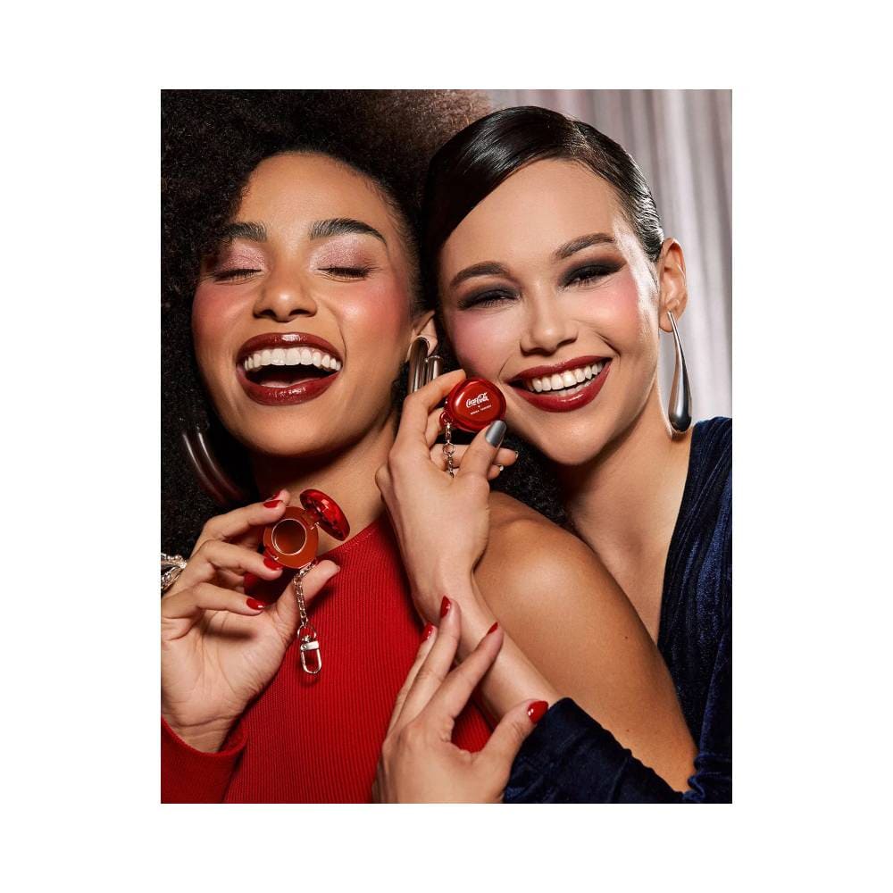 Duas mulheres segurando o Balm Labial Bruna Tavares Coca-Cola Pop Charm aberto mostrando o produto vermelho