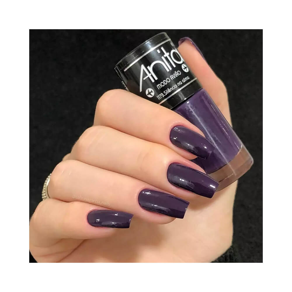 Mão segurando esmalte Anita Modo Avião Silêncio na Alma com unhas pintadas na cor roxa escura